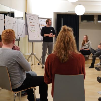 Alex steht am Flipchart vor der Runde der sitzenden Teilnehmenden und schaut ein Person aufmerksam an.